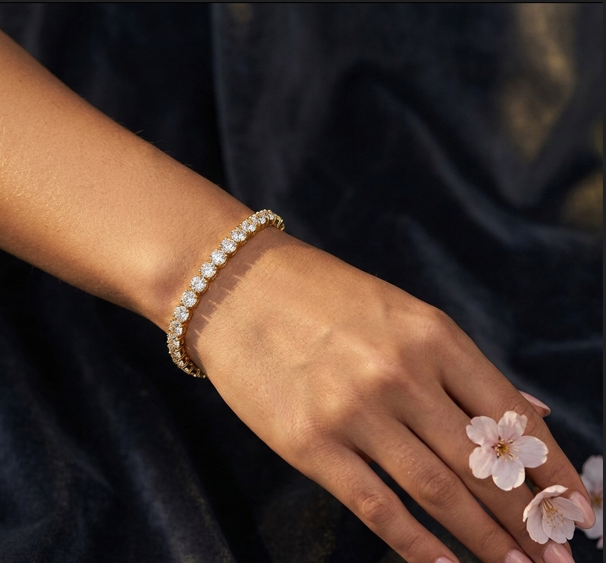 Hand wearing a gold bracelet with small diamonds, holding pink flowers against a dark background